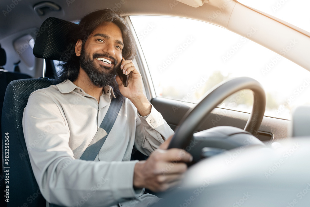A man with a beard enjoys driving his car while engaged in a phone conversation, demonstrating a relaxed and joyful mood under bright sunlight. A man with a beard enjoys driving his car while engaged in a phone conversation, demonstrating a relaxed and joyful mood under bright sunlight.