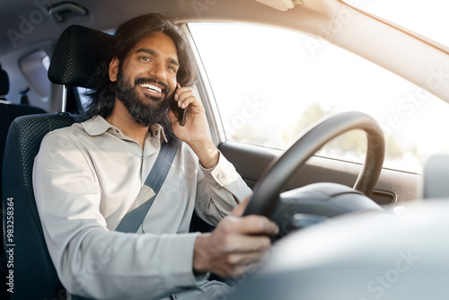 A man with a beard enjoys driving his car while engaged in a phone conversation, demonstrating a relaxed and joyful mood under bright sunlight. photo