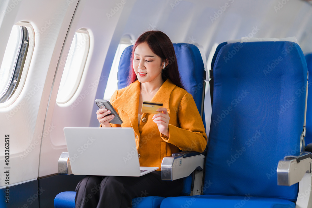 Businesswoman on a Flight: A smiling businesswoman in a yellow blazer sits on an airplane, using her phone, laptop, and credit card. The image captures the modern traveler, working on the go and stayi Businesswoman on a Flight: A smiling businesswoman in a yellow blazer sits on an airplane, using her phone, laptop, and credit card. The image captures the modern traveler, working on the go and stayi