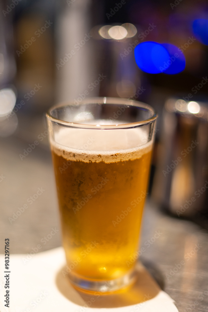 A clear pint drinking glass filled with cold foam and froth from a lager ale. The pale ale pint sits on the edge of a white metal patio table at a microbrewery with metal pub chairs in the background. A clear pint drinking glass filled with cold foam and froth from a lager ale. The pale ale pint sits on the edge of a white metal patio table at a microbrewery with metal pub chairs in the background.