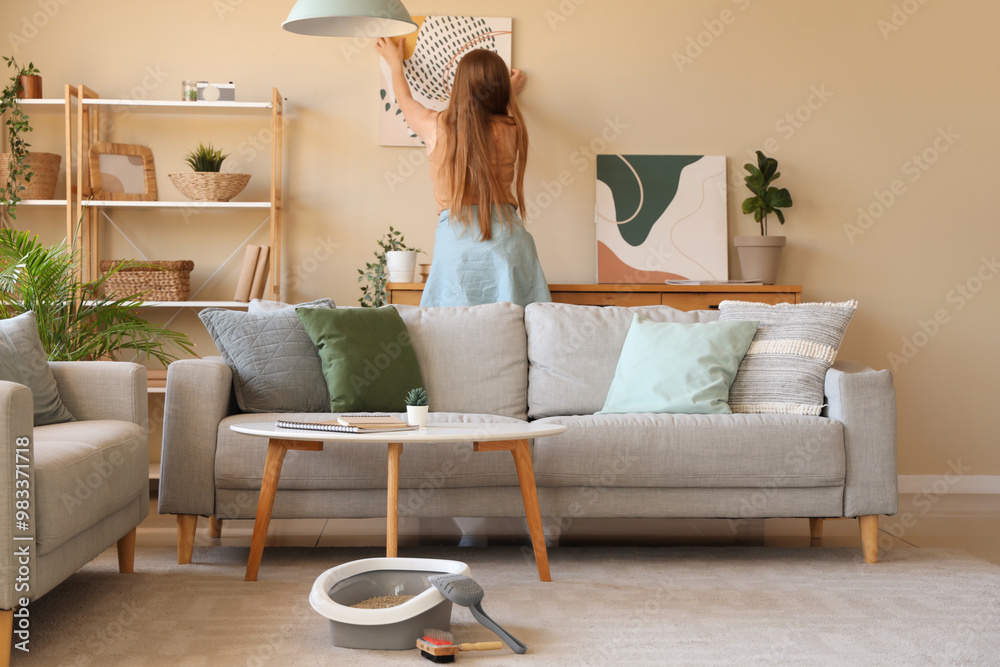 Young woman hanging painting on wall and cat litter box in living room