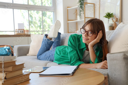 Smiling young woman in glasses relaxing on sofa with books and tablet, enjoyi...