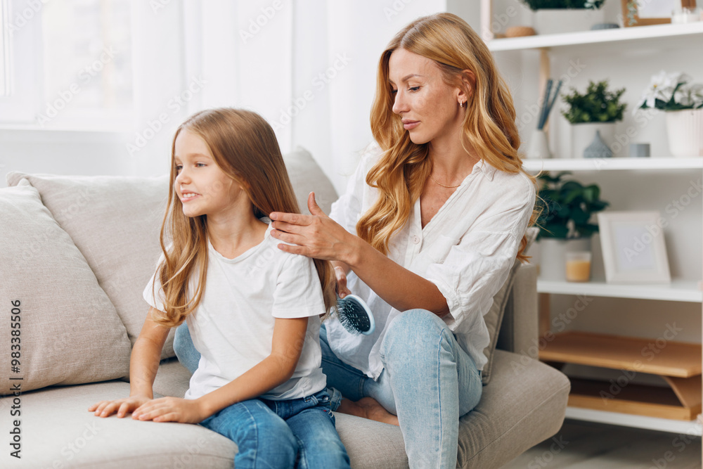 Mother combing daughter's hair while sitting on a cozy couch in a warmly lit living room setting Mother combing daughter's hair while sitting on a cozy couch in a warmly lit living room setting