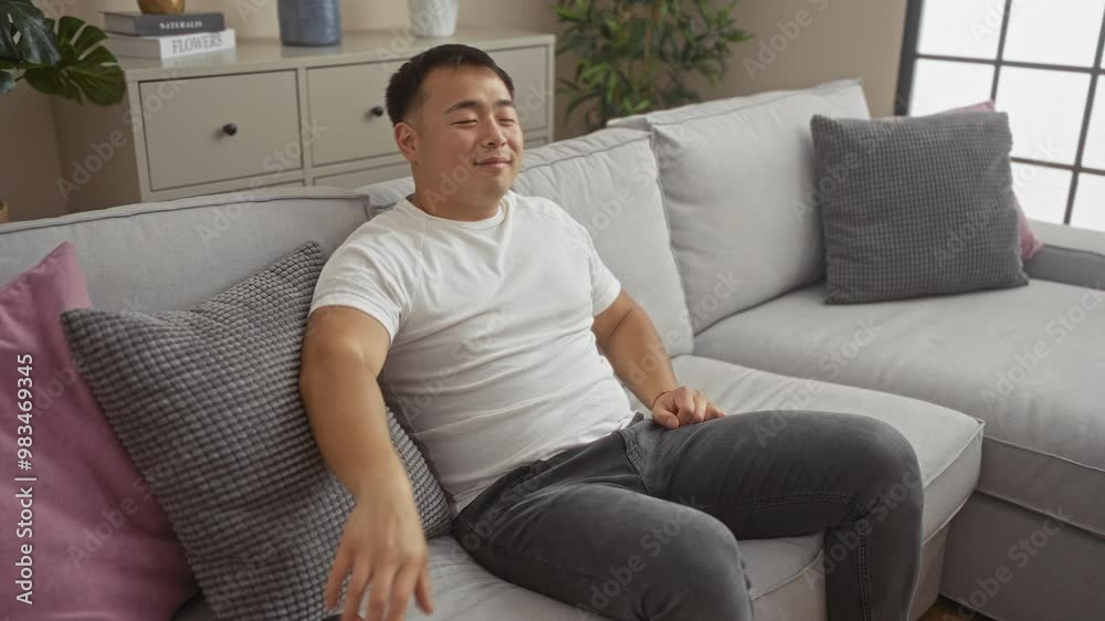A young asian man relaxes on a grey sofa in a modern living room, watching tv with a remote control in hand, enjoying leisure time at home.