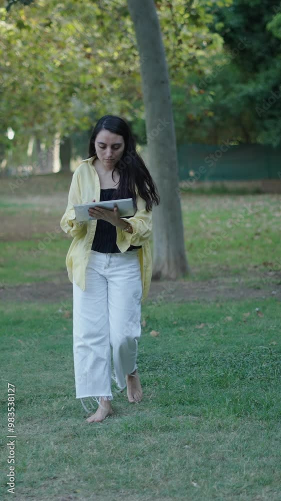 young brunette caucasian girl walking towards camera on the grass typing on the touch pad of a tablet young brunette caucasian girl walking towards camera on the grass typing on the touch pad of a tablet