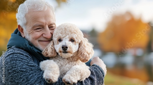 An elderly man holds a curly-haired dog tenderly outdoors, surrounded by autumn colors, reflecting calmness, companionship, and the warmth of friendships.