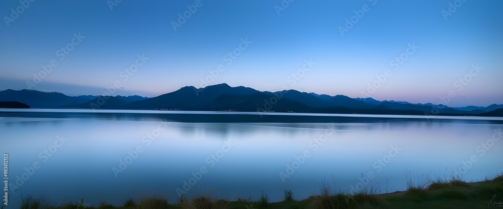 A landscape shot at dusk by the edge of a lake. Mountains on the horizon and the slightly blurred sky reflect on the calm surface of the water. A landscape shot at dusk by the edge of a lake. Mountains on the horizon and the slightly blurred sky reflect on the calm surface of the water.