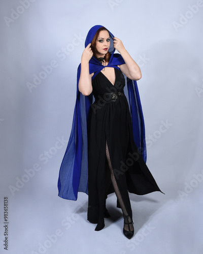 Full length portrait of  female red head model wearing  glamorous black witch dress with blue fantasy cloak halloween costume.  standing pose with gestural arms, isolated figure studio background.