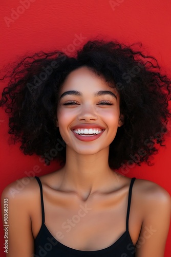 A young woman with curly hair smiles brightly at the camera. Her teeth are white and her eyes are closed. She is wearing a black tank top. The background is a solid red.