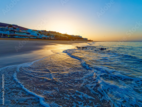 A beautiful sunrise over a sandy beach in Morro Jable on island Fourteventura. The sun is rising over a calm ocean, and the sky is a clear blue. There are palm trees and buildings in the distance.