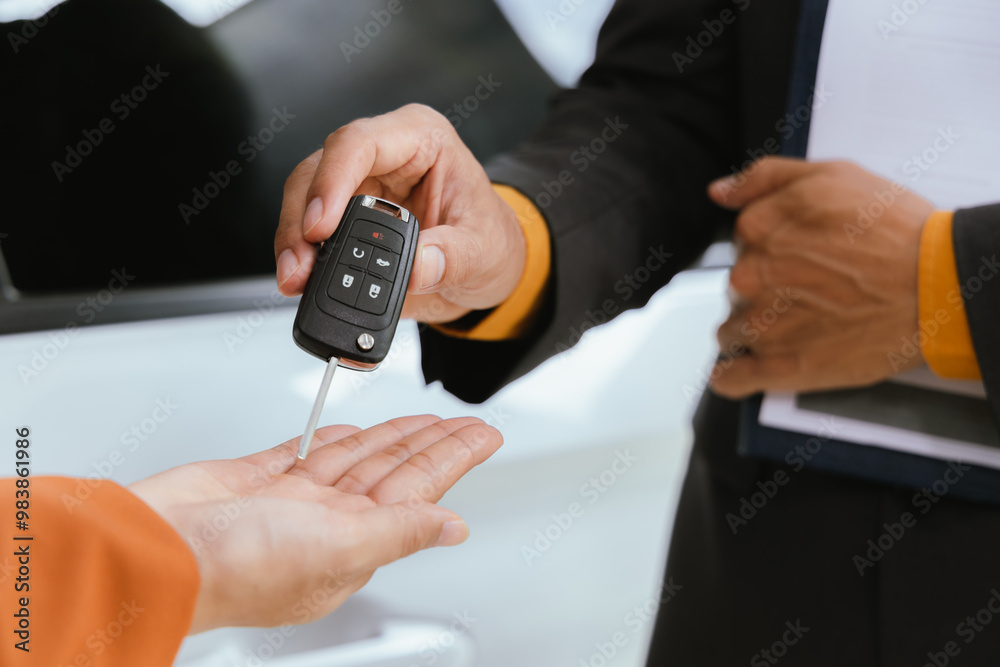 Fototapeta premium A businessman and his customer agree to a lease on a luxury car. The salesperson hands over the car keys as the customer signs a digital contract on a tablet to successfully close the sale.