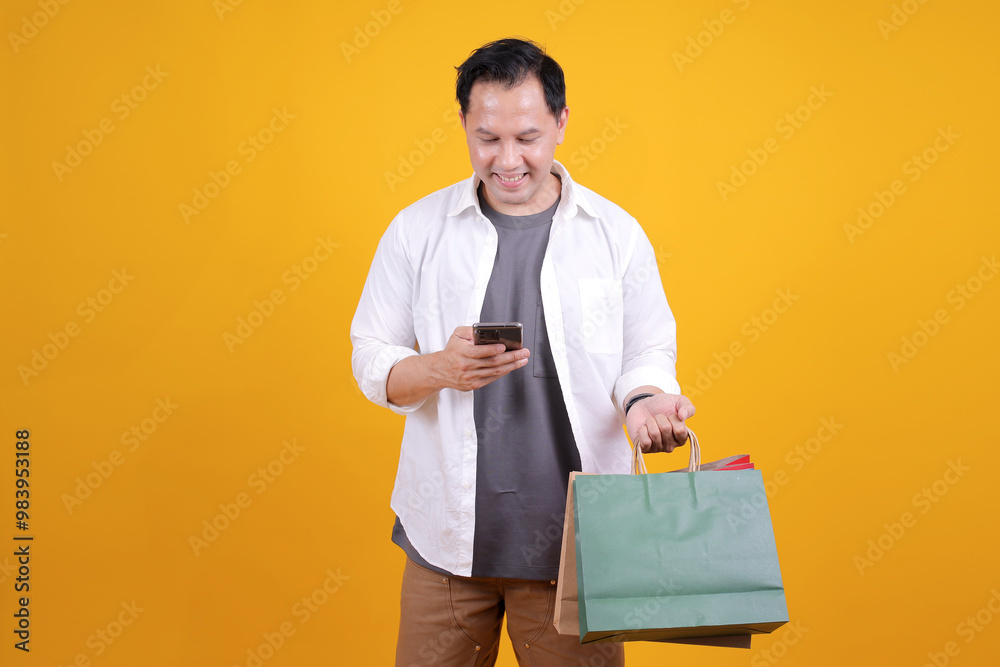 Happy Handsome Man Holding Shopping Bags While Using Smartphone Isolated on Yellow Background Happy Handsome Man Holding Shopping Bags While Using Smartphone Isolated on Yellow Background