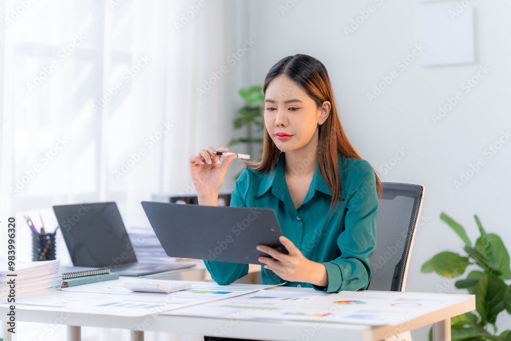 Focused Businesswoman Reviewing Documents at Desk: A young Asian businesswoman, dressed in a stylish teal blouse, sits at her desk, meticulously reviewing documents. She holds a pen thoughtfully, imme Focused Businesswoman Reviewing Documents at Desk: A young Asian businesswoman, dressed in a stylish teal blouse, sits at her desk, meticulously reviewing documents. She holds a pen thoughtfully, imme