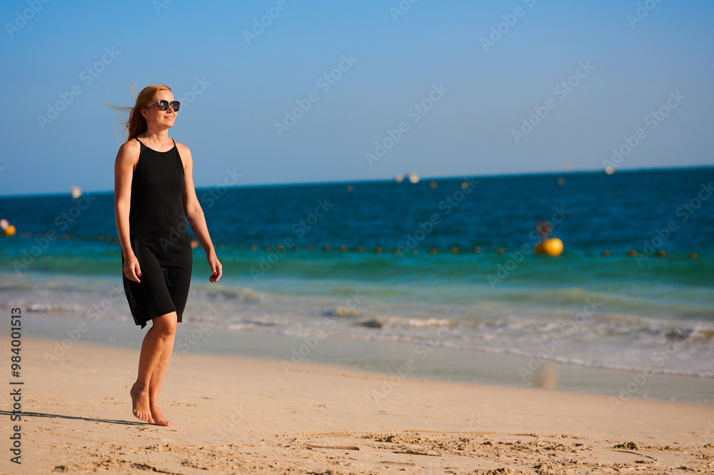 Spectacular blonde in black dress and sunglasses walks by the sea pleasantly spending her holiday. Spectacular blonde in black dress and sunglasses walks by the sea pleasantly spending her holiday.