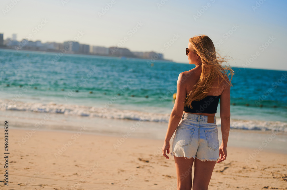 Young blonde European woman watches the sea waves from the back. A girl with a beautiful figure in shorts and top standing by the sea close-up. Young blonde European woman watches the sea waves from the back. A girl with a beautiful figure in shorts and top standing by the sea close-up.