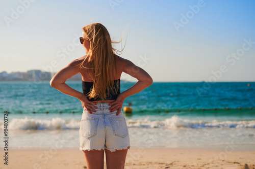 Blonde-haired girl stands on the coast. Back view of a Caucasian woman. Holiday, sea, sunset.