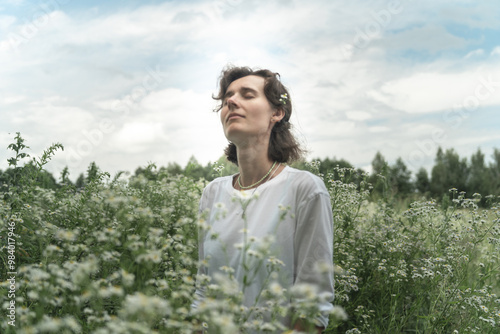 a girl of European appearance stands with her eyes closed in a chamomile field in a white T-shirt, blue sky, general plan