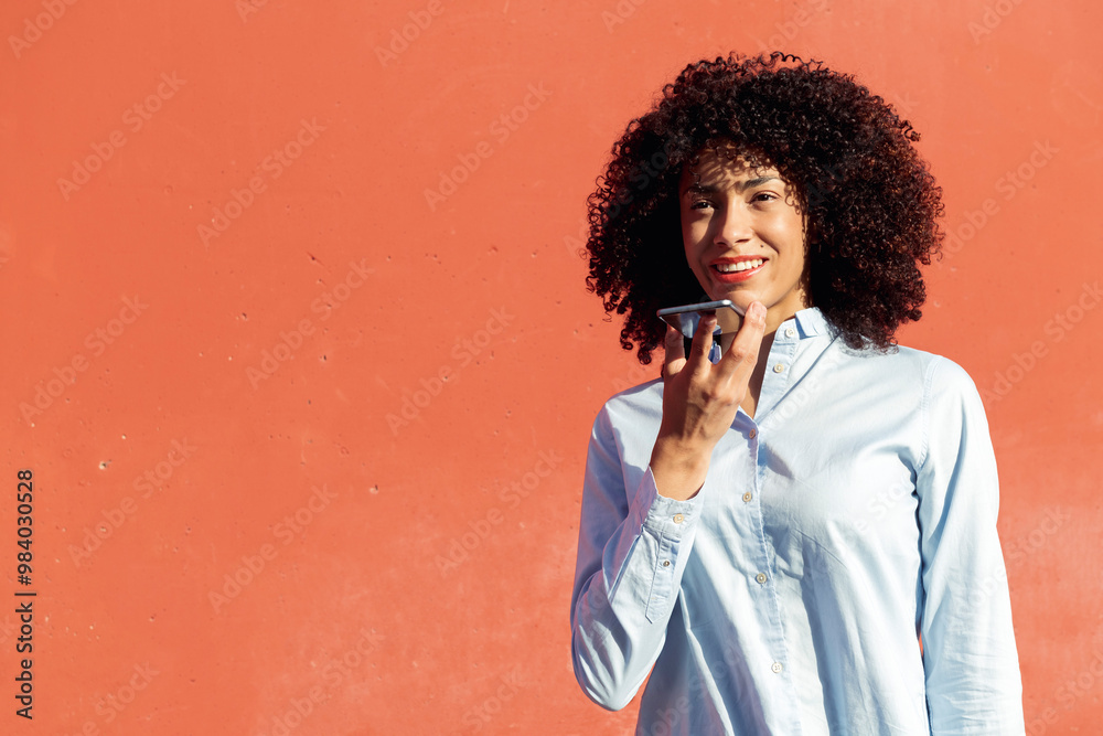 content ethnic female entrepreneur in formal shirt recording voice message on smartphone while standing in city on orange background