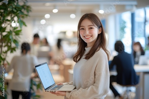 Smiling Japanese businesswoman standing in an office, holding her laptop. She is wearing casual clothes, and other people working behind her are blurred.,generated ai.
