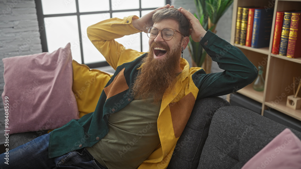 Fototapeta premium A cheerful bearded redhead man lounging on a couch indoors, expressing joy and comfort surrounded by books and plants.