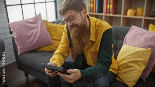 Smiling bearded man using tablet on cozy gray sofa with colorful pillows indoors