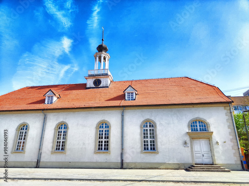 A white church with a red roof and a bell tower, surrounded by trees in a sunny, outdoor setting photo