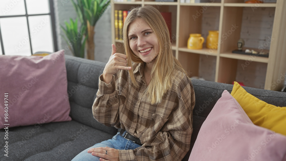 A smiling young blonde woman makes a call-me gesture while sitting on a couch indoors, surrounded by colorful pillows. A smiling young blonde woman makes a call-me gesture while sitting on a couch indoors, surrounded by colorful pillows.