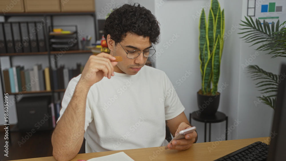 Fototapeta premium Handsome young hispanic man in office setting, holding phone and snack, sitting at desk indoors, with plants and shelves in the background.