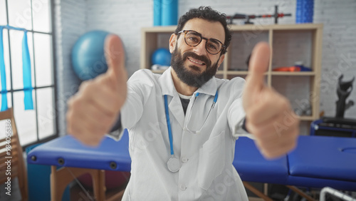 Wallpaper Mural Smiling bearded man in lab coat giving thumbs up in a modern clinic therapy room with rehabilitation equipment. Torontodigital.ca