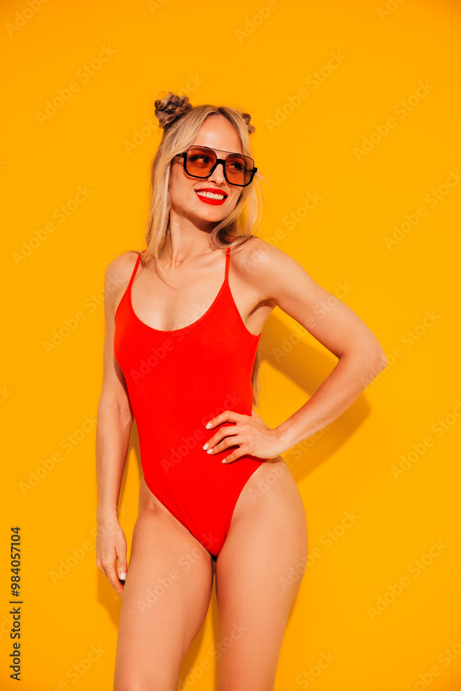 Young smiling blond model in summer swimwear colourful bathing suit. Sexy carefree woman having fun and going crazy. Female posing near yellow wall in studio. Cheerful and happy, sunglasses Young smiling blond model in summer swimwear colourful bathing suit. Sexy carefree woman having fun and going crazy. Female posing near yellow wall in studio. Cheerful and happy, sunglasses