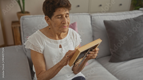 An elderly hispanic woman with short hair sits in her living room holding a picture frame, evoking emotions of nostalgia and reminiscence in a cozy home setting.