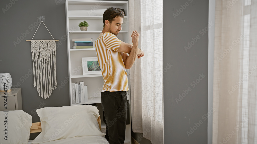 A young hispanic man in casual attire is standing thoughtfully in a well-lit, modern bedroom with stylish decor. A young hispanic man in casual attire is standing thoughtfully in a well-lit, modern bedroom with stylish decor.