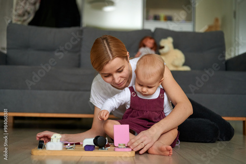 Girl of one and a half years old and her mother play an interesting game on the floor in the form of a board on which there is a switch, a socket with a plug, a lightning bolt, wheels and a cord.