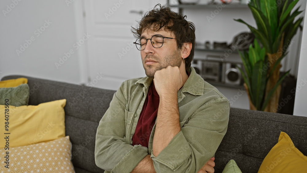 Obraz premium Pensive young hispanic man with beard and glasses sitting on a couch indoors with closed eyes