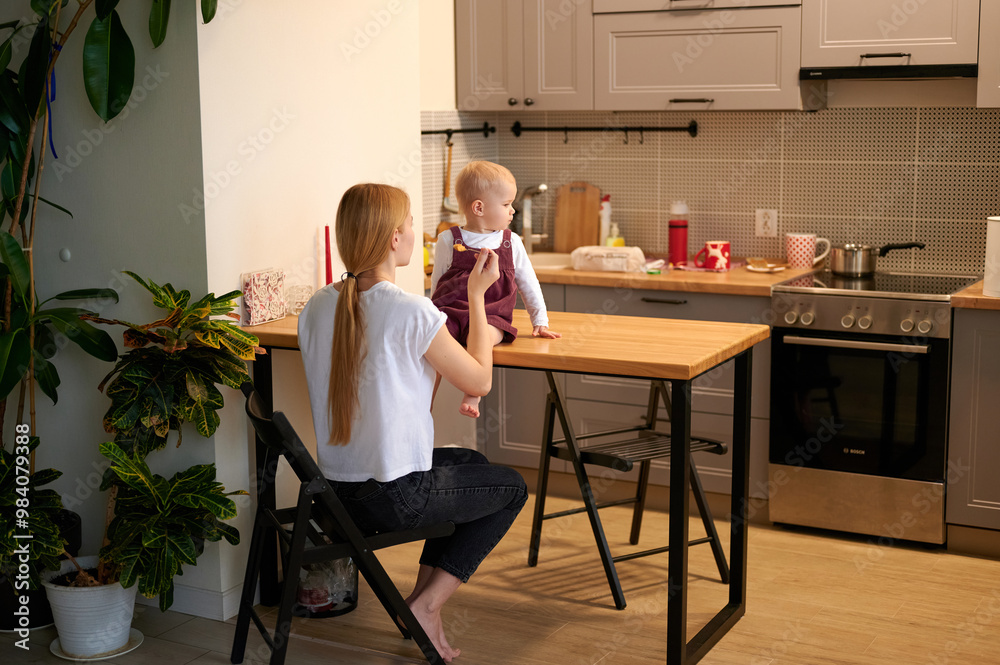 Mother feeding her baby. General view of the kitchen, mum sitting on a chair, little daughter on the table. Mother feeding her baby. General view of the kitchen, mum sitting on a chair, little daughter on the table.