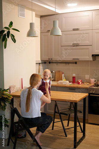 Mother feeding baby daughter. General view of the kitchen, mum sitting on a chair, little daughter on the table. A mum and a girl in white food shirts.