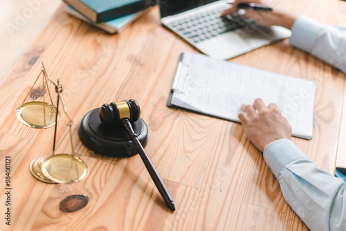 A person writes notes while using a laptop beside a gavel and scales of justice, symbolizing legal work or law practice.