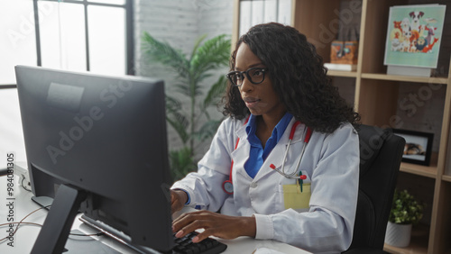 A young african american female doctor with curly hair works on a computer in a clinic office, displaying focused intent and professional demeanor in a modern, well-lit medical workplace.