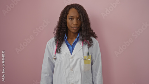 Young african american female doctor with curly hair in a white coat against an isolated pink background.