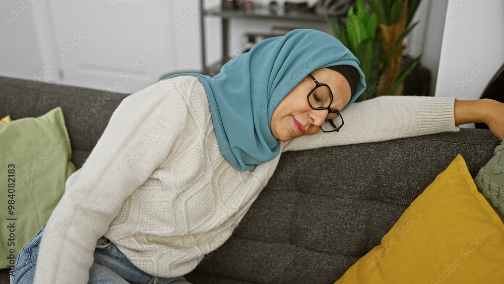 A serene middle-aged woman with glasses and a hijab resting peacefully on a couch at home. A serene middle-aged woman with glasses and a hijab resting peacefully on a couch at home.