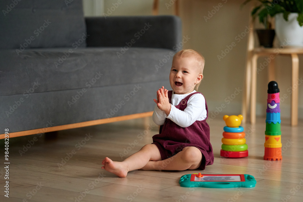 Beautiful blonde-haired girl and a bright magnetic board, with coloured plastic pyramids nearby. Happy child sits on the floor with his toys and claps his hands. Beautiful blonde-haired girl and a bright magnetic board, with coloured plastic pyramids nearby. Happy child sits on the floor with his toys and claps his hands.