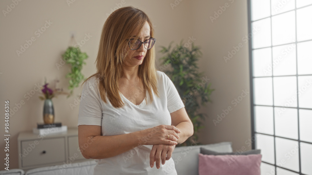 Middle-aged woman with blonde hair wearing glasses and a white shirt standing in a bright, cozy living room interior. Middle-aged woman with blonde hair wearing glasses and a white shirt standing in a bright, cozy living room interior.