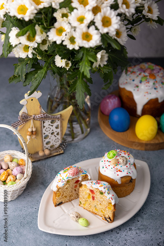 Two small Easter kulichs with candied fruits in white glaze with colorful sprinkles in the cut. Painted chicken and quail eggs. Traditional Easter baking. Easter holiday. Close-up, selective focus.