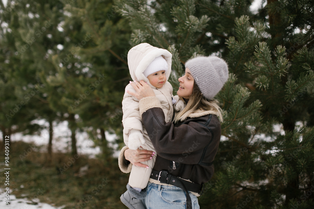 Woman in a brown short sheepskin coat is holding a baby dressed warmly in a white overall and hat, surrounded by snow-covered ground and evergreen trees. Joyful moment between mom and baby. Woman in a brown short sheepskin coat is holding a baby dressed warmly in a white overall and hat, surrounded by snow-covered ground and evergreen trees. Joyful moment between mom and baby.