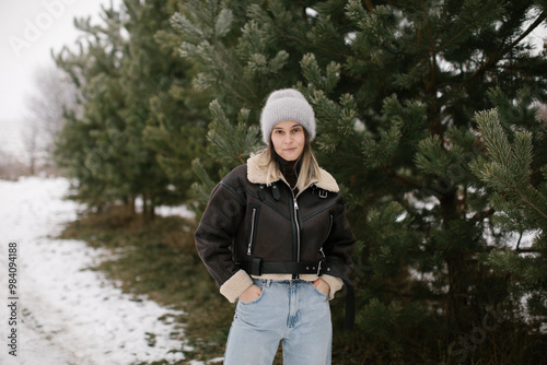 A woman in a short brown sheepskin coat and jeans stands near evergreen trees in winter. photo