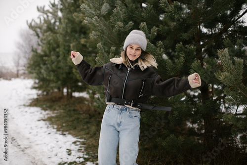 A woman in a short brown sheepskin coat and jeans stands near evergreen trees in winter. Woman dressed warmly is looking up. photo