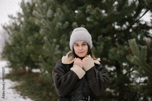 A woman in a short brown sheepskin coat and jeans stands near evergreen trees in winter. photo