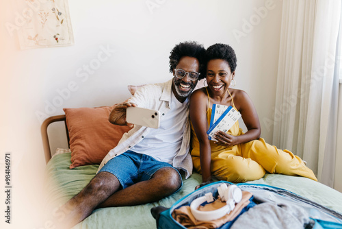 Excited couple smiling on bed with flight tickets, making a video call before a romantic getaway