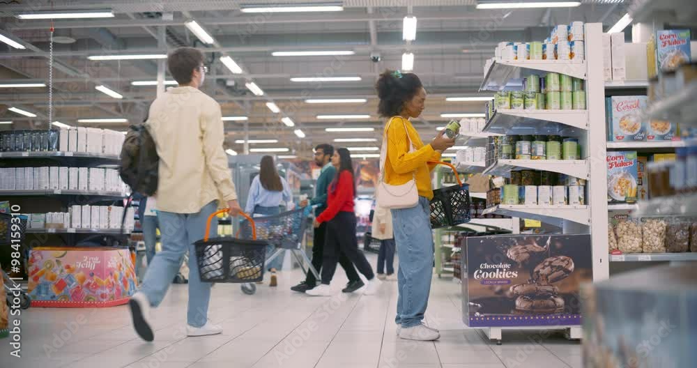 Young Black Female in Yellow Sweater Shops for Groceries in a Supermarket, Browsing Shelves and Selecting Items. Other Shoppers with Baskets and Carts are Visible in the Background Young Black Female in Yellow Sweater Shops for Groceries in a Supermarket, Browsing Shelves and Selecting Items. Other Shoppers with Baskets and Carts are Visible in the Background