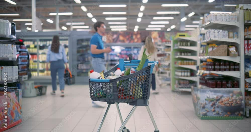 Establishing Footage with a Shopping Cart Standing Between the Aisles in a Modern Grocery Store. Busy Supermarket with Diverse People in the Background. Trolley with Vegetables, Milk, Canned Foods